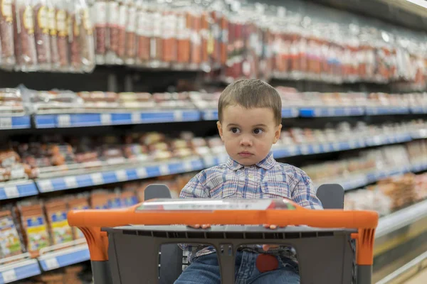 Um menino bonito vestido com uma camisa xadrez em um supermercado em um carrinho. Contra o pano de fundo de prateleiras com produtos à base de carne e salsichas . — Fotografia de Stock