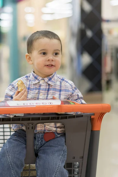 Щаслива дитина в сорочці сидить у візку в торговому центрі і їсть хліб — стокове фото