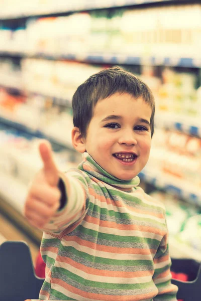 Boldog fiú egy szupermarketben a polcok mögött, étellel. A fiú megmutatta, hogy minden rendben. függőleges fénykép — Stock Fotó