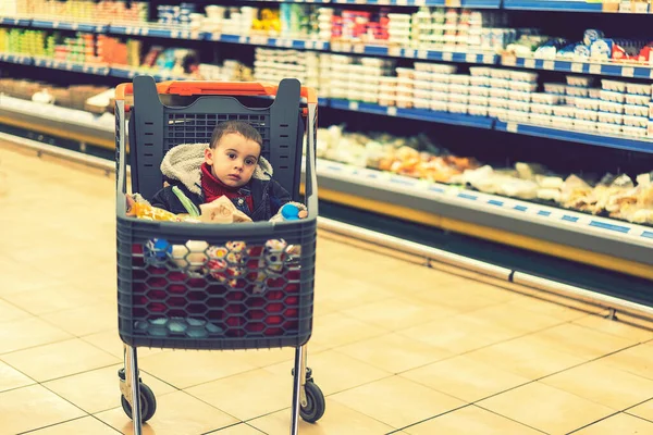 Hermoso bebé en el carrito de la compra - carro. Un carro con productos en el que se sienta el niño. Carro completo con comida en el supermercado. En el carro se sienta un bebé. tonificado — Foto de Stock