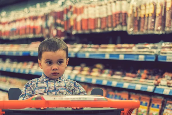 Симпатичный мальчик, одетый в клетчатую рубашку в супермаркете на тележке. На фоне полок с мясными продуктами и сосисками. тонированные — стоковое фото