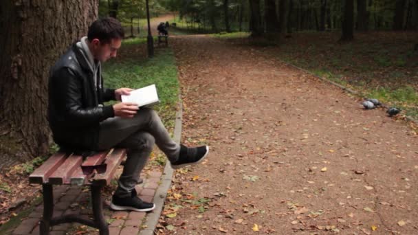 Чоловік читає книгу, сидячи на лавці — стокове відео