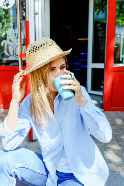 Closeup düşünceli fincan kapuçino tutan kot görünümlü kız. Sarı saçlı ve hasır şapka. Yaşam tarzı kadın portre