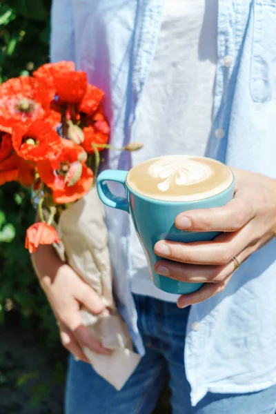 Blonde girl holding poppy flowers bouquet and a blue cup of warm morning cappucino. Lifestyle hipster portrait
