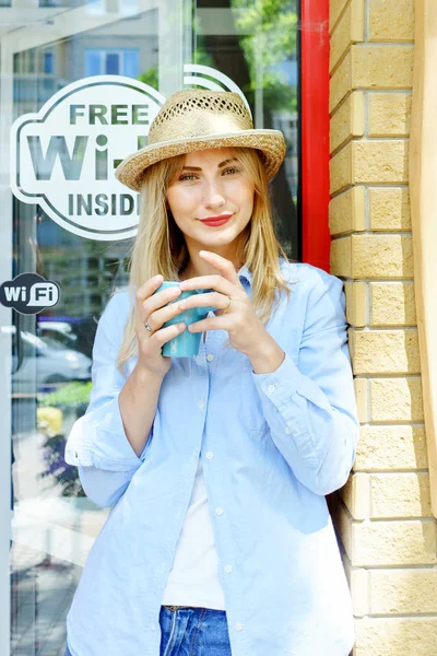 Sarışın genç kız kamera arıyorum. Kot pantolon ve hasır şapka giymek. Fincan kapuçino holding