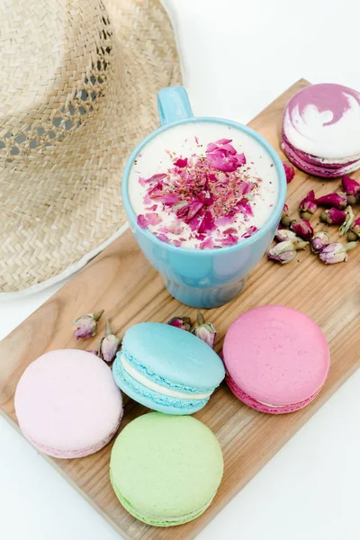 Gustosi biscotti macaron con una tazza blu di cappuccino con petali di rose e cappello di paglia sul tavolo bianco . — Foto Stock