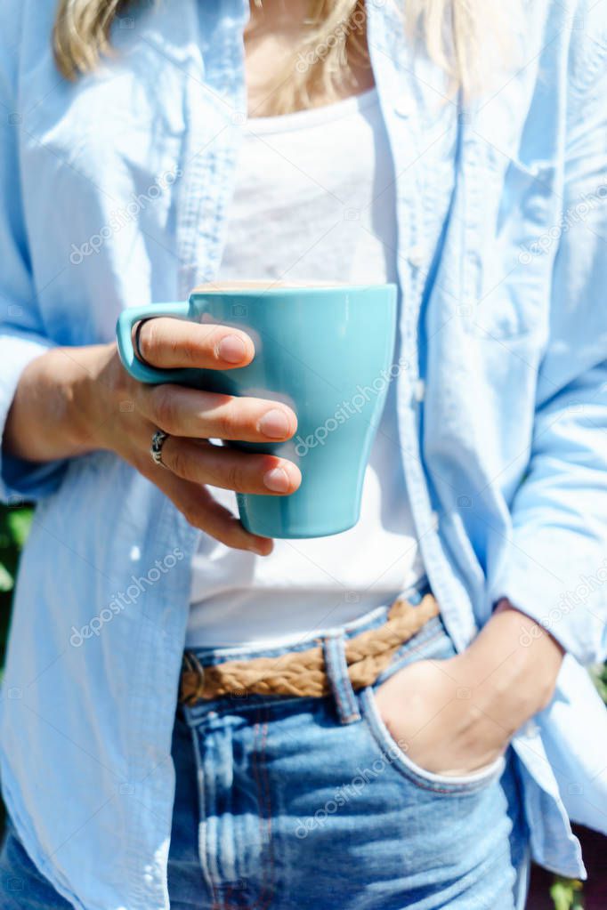 Young female hipster person in jeans wear holding a blue cup of warm cappuccino