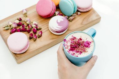 Kadın elini tut kapuçino Kupası gül yaprakları ile. Güzellik Fransız macarons ahşap masa ve beyaz arka plan üzerinde