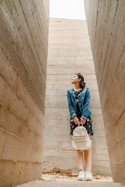 Attractive young woman stands between concrete walls. Caucasian girl in casual jeans and flower dress looks up.