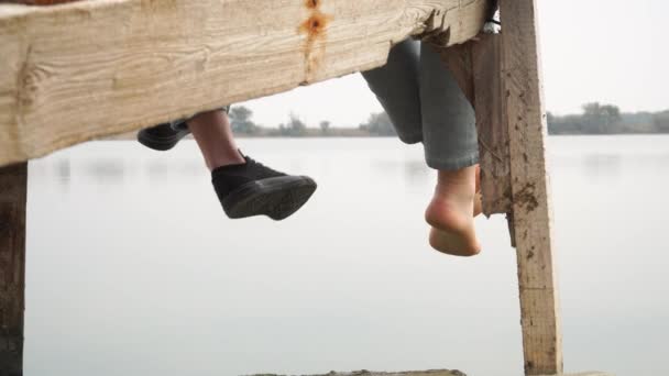 Dwie dziewczyny relaksujący siedzący na drewniane molo nad jeziorem huśtawka stóp — Wideo stockowe