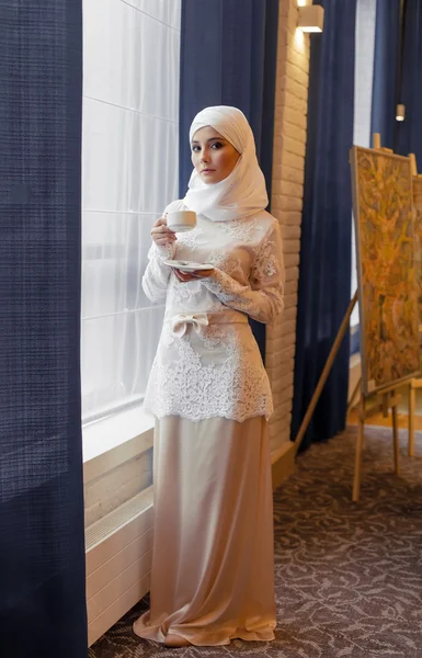 Wanita muslim cantik dalam gaun pengantin putih berdiri di jendela — Stok Foto