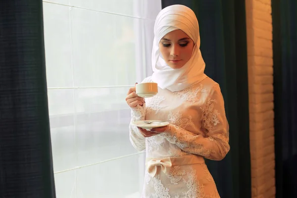 Wanita muslim cantik dengan gaun pengantin putih dengan secangkir teh di tangannya — Stok Foto