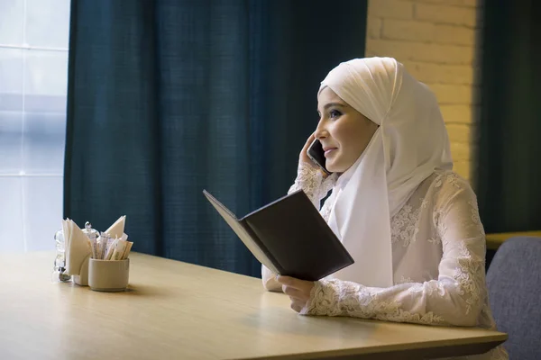 Muslimsk kvinna i en vit klänning som pratar i telefon vid bordet på ett kafé — Stockfoto