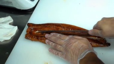 Şeffaf steril eldivenli Asyalı adam mutfaktaki beyaz bir masada satılık keskin bir bıçakla ustalıkla kızarmış balığın yarısını kesti. Üst Manzara. Suşi ustası lezzetli ekmekler yapıyor..