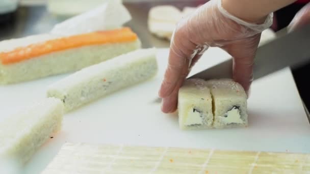 寿司主菜卷 一个戴透明无菌手套的男人巧妙地在厨房的白桌子上用一把锋利的小刀切着面包卷 饭和鱼卷准备晚饭 — 图库视频影像