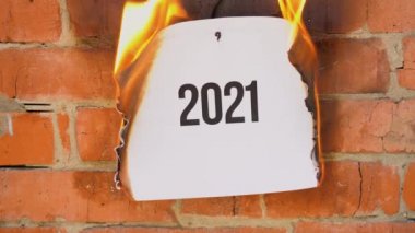 Beyaz bir A4 tablosundaki yazı turuncu bir duvarın arka planına karşı. Üzerinde 2021 yazan kağıt yanıyor, duman çıkıyor ve küle dönüşüyor..