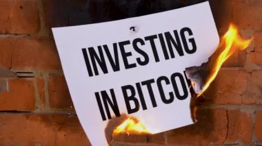 Beyaz bir A4 tablosundaki yazı turuncu bir duvarın arka planına karşı. Bitcoin 'e Yatırım' ın siyah sözcüğünün olduğu gazete yanıyor, duman çıkıyor ve küle dönüşüyor. İş konsepti.