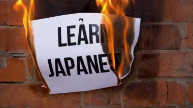 Beyaz bir A4 tablosundaki yazı turuncu bir duvarın arka planına karşı. Üzerinde siyah kelime olan gazete Japonca yanıkları öğreniyor, sigara içiyor ve küle dönüşüyor. İş konsepti.