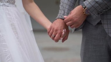Mutlu evli çiftin el ele tutuşması. Kareli takım elbiseli yakışıklı damat ceketinin kollarını düzeltiyor ve açık havada beyaz gelinlik içinde genç gelinin elini tutuyor..