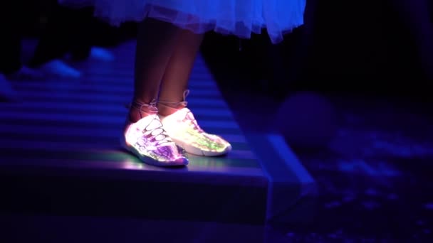 一个穿着时髦的白色发光运动鞋和裙子的女孩的腿在一个黑暗的俱乐部的舞台上与许多人亲密接触 那个女孩呆在现场跳舞 鞋子的光照亮了 — 图库视频影像
