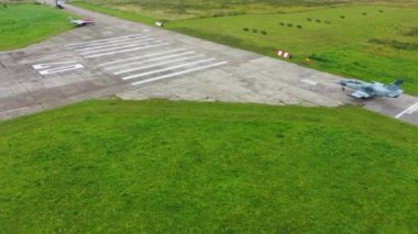 Gri mavi bir savaş uçağı yeşil alanlar arasındaki bulutlu bir günde eski havalimanındaki piste doğru yola çıkıyor. İHA 'nın üst görüntüsü. Uçak beton levhalar boyunca 7 numaraya hareket ediyor..