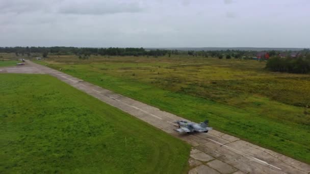 灰色の青い戦闘機は緑のフィールドの間で曇りの日に古い空の飛行場の滑走路に沿って移動します 四角形の上からの眺め 飛行機はコンクリートスラブ上を移動します — ストック動画