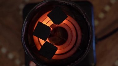 Kömürler, yürüyüş için eski ve özel bir elektrikli ocakta tutuşturulur ve yakılır. Üç siyah kare kömür kırmızı-sıcak turuncu bir spiral üzerinde uzanır ve yanar yakın plan bulanık kahverengi. Yukarıdan görüntüle