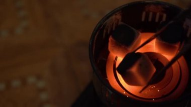 Kömürler eski elektrikli kamp sobasında yanıyor ve yanıyor. Metal maşalı bir adam, sağında bulanık bir arkaplanda kızgın bir spiral üzerinde 3 siyah kare kömür atar. Üst Manzara. Nargile kavramı