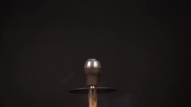 Güzel bir bej nargileden beyaz duman dumanı yükselirken siyah arka planda esiyor. Kahverengi bir kase ve metal bir kaloud nargileye konur..
