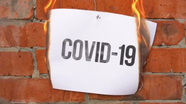 Beyaz bir A4 tablosundaki yazı turuncu bir duvarın arka planına karşı. Üzerinde COVID-19 yazan kağıt yanıyor, tütüyor ve küle dönüşüyor. Dünya Savaşı konsepti.