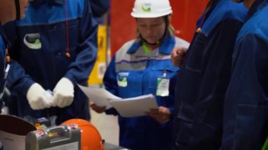 Sochi, Rusya - 07 Ağustos 2019: Müfettiş kadın patron mavi koruyucu elbise giyiyor, beyaz kask takıyor, kağıtları elinde tutuyor, insanlara bakıyor, fabrikada tamamlanmış mühendisler grubunun çalışmalarını yakından kontrol ediyor. Arka taraftaki yazı - katılımcı