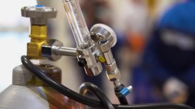 Large metal gas cylinder with a glass tube, inside scale with numbers. Gearbox is properly connected to down going hose. Valve is tightened close up. People walks on blurred background of factory.