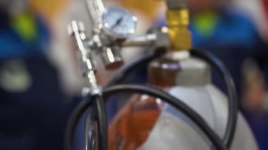 Vites kutusuna bağlı metal gaz balonu mükemmel durumda, valf sıkıca sıkıyor. Uzun kauçuk bir hortum iniyor. Bulanık arka planda insanlar ayakta duruyor, tulum giyiyor, kask takıyor.