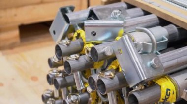 Gri yeni metal boruların yakın görüntüsü, fabrikanın atölye alanında detaylar yığılmış durumda. En üstte, tahta palette kesilmiş demirler var. Arka plandaki mavi pantolonlu adam geçerken.
