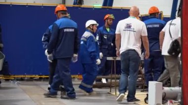 Sochi, Russia - August 07 2019: In room, many workers people wear special clothing, helmets, glasses on background of blue screens. Employees men discuss the work plan. On floor lay tools and bundles of fittings. Indoors.