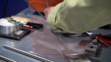 Kaynakçı ellerinin dar koruyucu bornozlu görüntüsünü kapat. Tanınmayan adam depodaki metal tezgahtan kesilmiş tel parçaları alıyor. Kablolu kaynak makinesi, mengene, masanın üzerinde duran diğer araçlar.
