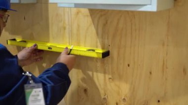 Beyaz işçi mavi takım giyer, kalem ve inşaat seviyesinde kontrplak kaplamayla çizer. Adamın üstünde iki beyaz metal kutu var zaten..