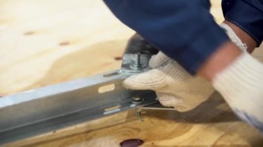 Birkaç parçadan oluşan işçi kolu, yerde duran metal rafa iki cıvatalı ve yakın konteynırlı plaka takıyor. Adam beyaz koruyucu eldiven ve iş kıyafeti giyiyor..