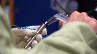 Erkek kaynakçı koruyucu bornoz giyer, takım elbise giyer, duvar kalınlığını ölçer, metal gri yontulmuş parça, özel cetvelle kesilmiş tüp parçası. Adam sağ elinde ölçüm aletini tutuyor, kalibre yakın..