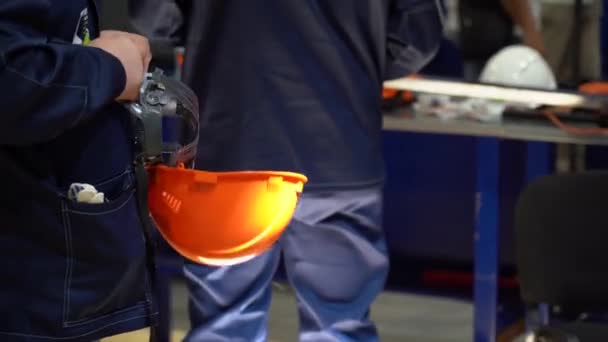 労働者のグループは 生産現場を背景としたツールで作業台の近くに立っている 男は青い特別な制服を着ている 従業員はオレンジ安全ヘルメット 眼鏡を保持し 頭のためのサイズを調整します — ストック動画