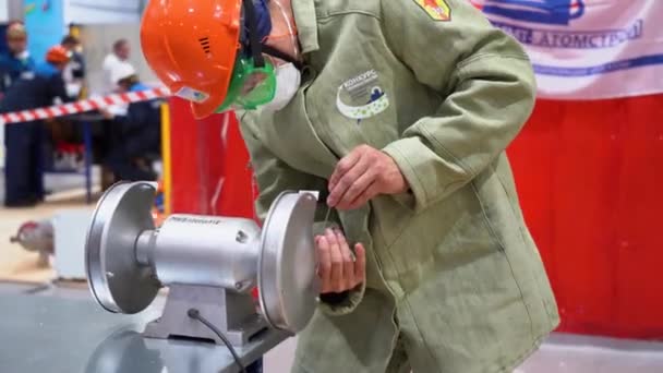 俄罗斯索契 2019年8月7日 白种人工人在工厂背景下在工作场所的磨床上研磨金属棒 一名男子身穿特殊的绿色防护服 戴着白色的面罩 头上戴着闪烁着的红色头盔 — 图库视频影像