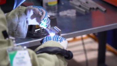 Tulumlu kaynakçı, beyaz koruyucu eldiven giyiyor yarı otomatik bir makineyle kaynak yapıyor. Atölye mengeneye sıkıştırıldı. Adam tezgahın dikişine metal ekliyor, fabrikanın manzarasını kapatıyor.