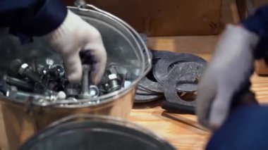 İşçi ellerinin yakın görüntüsü. Mavi iş elbiseli iki adam, eldivenler tahta zemindeki çelik kovalardan vida somunlarıyla benzer metal cıvatalar seçiyorlar. İçeride, endüstri. Fabrikada günlük çalışma.