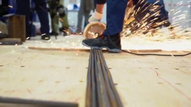 Erkek işçi demir demirin kenarını demirlere, ütülere, elektrikli testerelere ve öğütücüye dayamış. Kıvılcımlar farklı yönlerde uçar. Birçok insan atölyeye fabrika arka planında yürür. Uzun çubuklar tahta zeminde yatıyor..