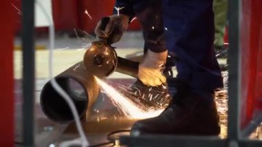 Tanımlanamayan profesyonel müteahhit mavi üniforma, beyaz koruyucu eldiven, siyah çizme, yakın plan. Erkek elinde öğütücü, büyük metal borular kesiyor. Kıvılcımlar uçar. Kamera yakınlaştırılıyor.