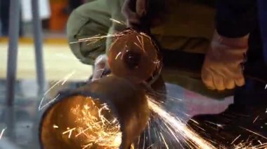 Erkek çalışan koruyucu mavi üniforma giyiyor, ellerinde beyaz eldivenler. Adam paslı metal boruyu öğütücüyle kesiyor. Kıvılcımlar her yöne uçar. Bir sonraki işçi yardım et, tut, boruyu döndür..