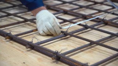 Beyaz koruyucu eldiven giyen, mavi ceketli, elinde örgü kablosu tutan, demir çubuklar arasındaki mesafeyi ölçen bir erkek işçi. Adam ahşap zeminde metal bir çerçeve hazırlıyor..