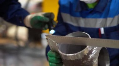 İki işçi yeşil eldiven, koruyucu tıbbi maske ve gözlük takıyor. Adam elinde küçük bir metal boru parçası, kenarları soyulmuş, ölçüleri köşeli cetvelle ölçülmüş, kontrolleri düzgün, çapı fabrikada.. 