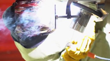 Male construction worker wear green jacket, protective helmet, yellow gloves performs work, use welding machine. At point of contact electrode with metal surface, bright glow, sparks fly out close up