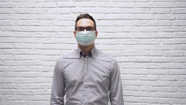 灰色のシャツを着た黒い眼鏡のハンサムな白人男性実業家は 彼の顔から保護ターコイズの医療用マスクを脱ぎ 白いレンガの壁の背景に笑顔 癒しのコンセプト — ストック動画